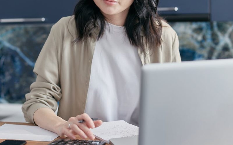 A young woman calculating finances at home using a laptop and calculator, reflecting a focused budgeting process.