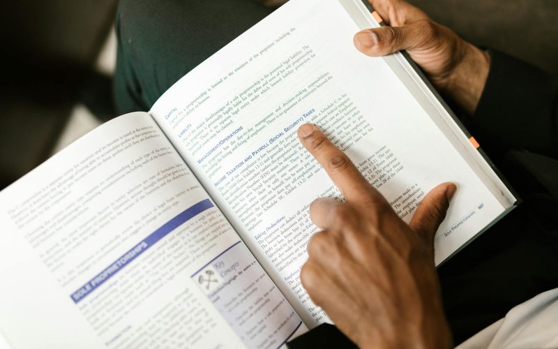 An adult reading a book on taxes, focusing on specific text with a pointing finger.