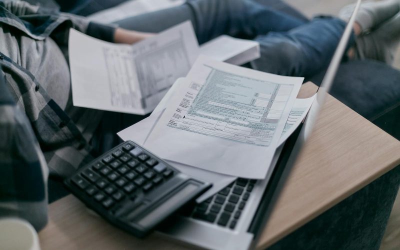 A young adult using a calculator and laptop to manage finances at home, surrounded by papers.